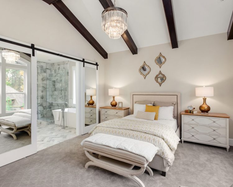 Elegant master bedroom featuring a vaulted ceiling with dark wood beams, a crystal chandelier, mirrored barn doors opening to an ensuite bathroom, upholstered bed with nightstands and lamps, and plush gray carpeting.