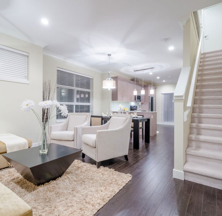 Bright finished basement featuring an open concept living area with beige sectional sofa, white accent chairs, dark hardwood flooring, shag area rug, modern coffee table with flowers, carpeted staircase, recessed ceiling lights, pendant lighting, and a kitchen area visible in the background with light wood cabinetry