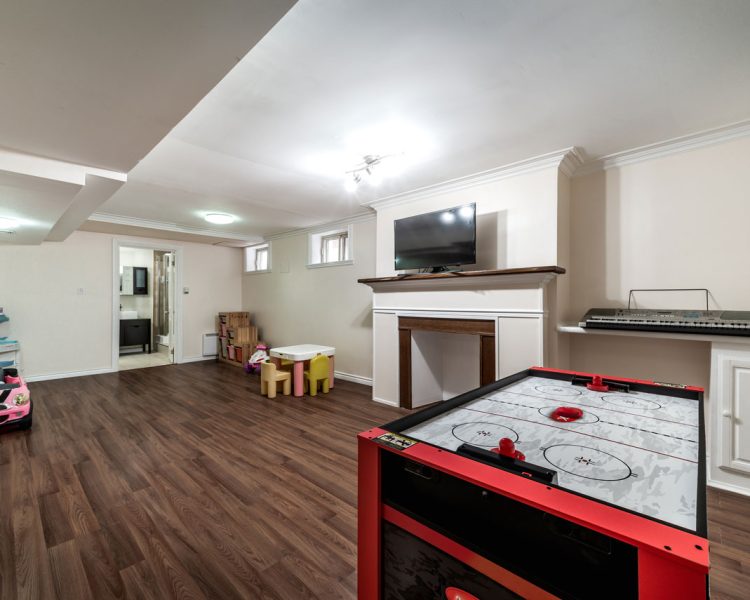 Spacious basement playroom featuring dark wood laminate flooring, red air hockey table, pink toy car, wall-mounted TV above fireplace mantle, electronic keyboard on built-in shelf, children's table and chairs, and recessed ceiling lights throughout