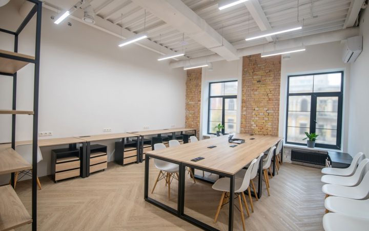 Modern office interior with light wood flooring, long wooden conference tables with white Eames-style chairs, exposed brick accent wall, industrial white ceiling with beam details, large windows, and minimalist black shelving units