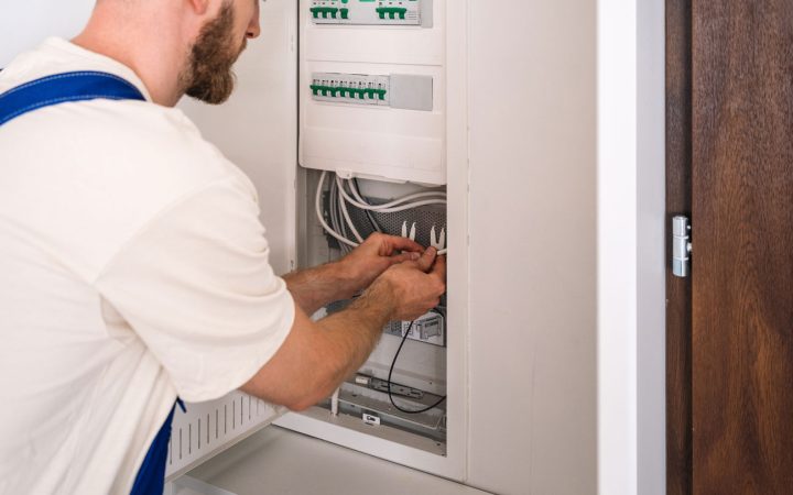 Skilled electrician wearing white shirt and blue suspenders organizing and connecting wiring inside an open electrical breaker panel with green terminal blocks and organized cable management
