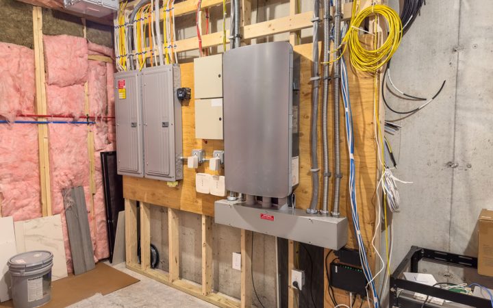 An extensive residential electrical setup in an unfinished basement featuring multiple breaker panels, a large inverter or battery backup system, and organized conduit wiring against exposed studs and insulation.
