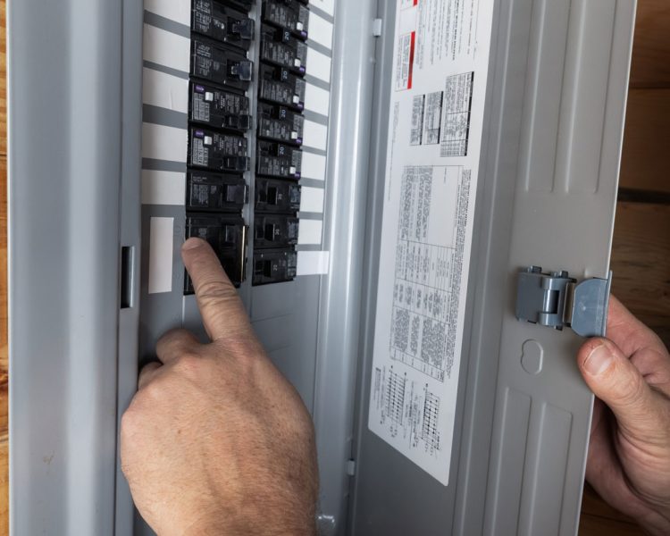 A close-up view of an electrician's hand flipping a switch on a circuit breaker inside a gray electrical panel box mounted on a wood-paneled wall.