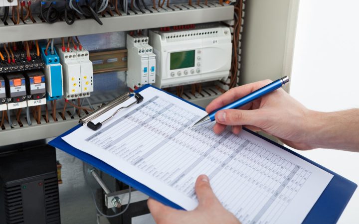 A technician's hands holding a clipboard and pen, reviewing a checklist in front of an open industrial electrical control panel containing programmable logic controllers (PLCs), contactors, and complex wiring.
