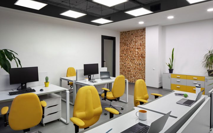 Bright modern office workspace with white desks, vibrant yellow ergonomic chairs, black exposed ceiling with square LED panel lights, exposed brick accent wall, computer workstations, and indoor plants