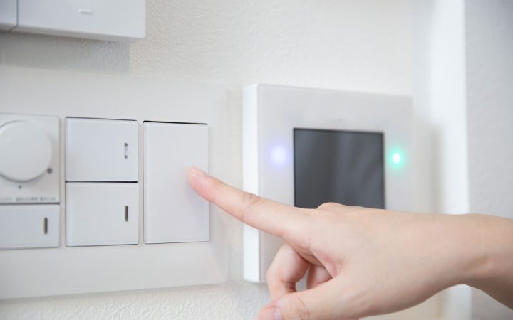 Close-up of a person's finger pressing a modern white rocker light switch next to a digital smart home control panel with illuminated blue and green indicator lights mounted on a white wall