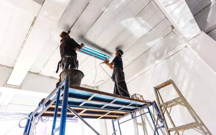 Two electricians standing on blue scaffolding installing or servicing fluorescent light fixtures in a high-ceiling commercial building, demonstrating professional commercial electrical contracting services