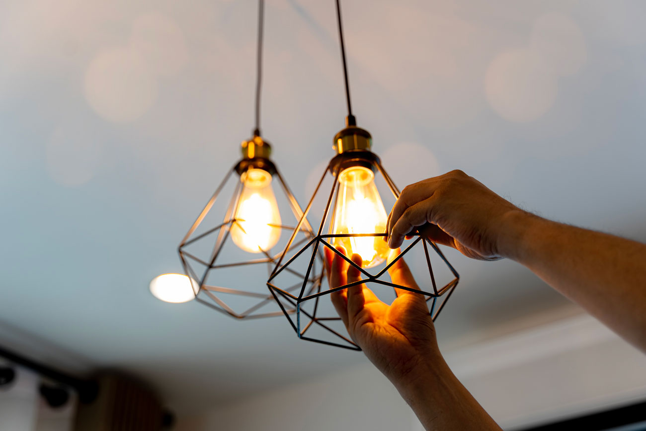 Close-up of hands installing or adjusting contemporary black wire geometric cage pendant lights with warm glowing Edison bulbs.