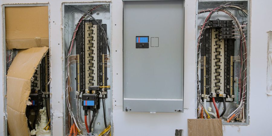 Wall-mounted electrical breaker panels during installation, showing exposed wiring and circuit breakers in open boxes alongside a closed gray metal panel cover.