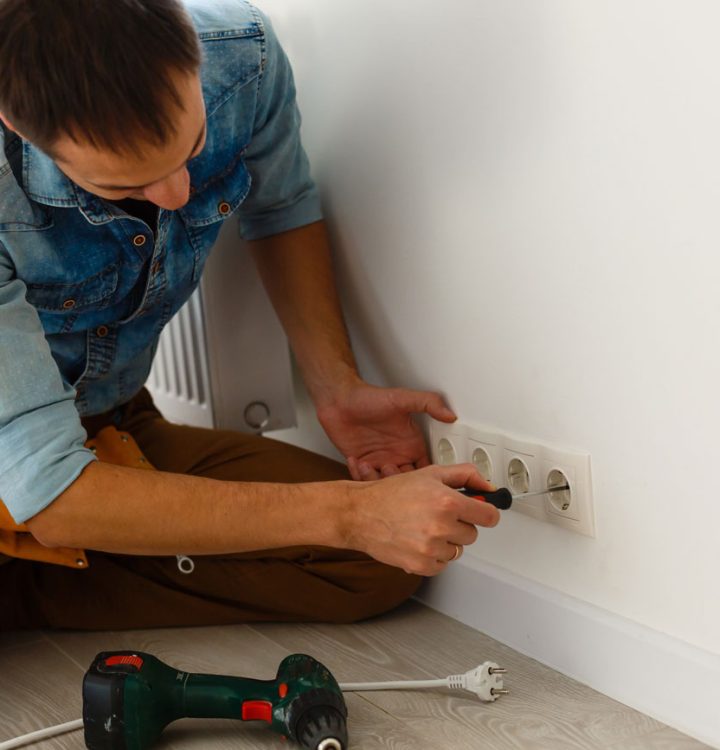 A handyman in a denim shirt kneels on the floor, using a screwdriver to install a series of white European-style electrical outlets on a clean white wall.