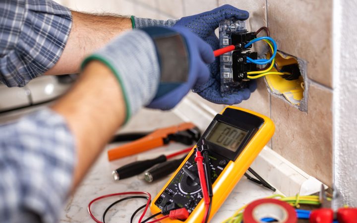An electrician wearing blue gloves uses a multimeter to test the wiring of an exposed electrical outlet, with various tools like pliers and wire cutters on the marble countertop.