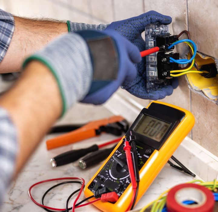 An electrician wearing blue gloves uses a multimeter to test the wiring of an exposed electrical outlet, with various tools like pliers and wire cutters on the marble countertop.