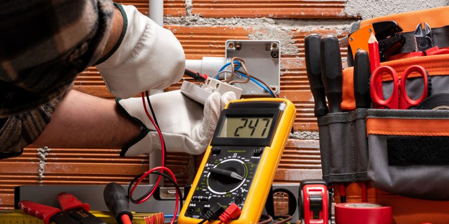 Electrician wearing white protective gloves using a yellow digital multimeter displaying 24.7 volts while working on electrical wiring in an exposed junction box, surrounded by professional tools including wire strippers, screwdrivers, and an orange tool bag against a brick wall background