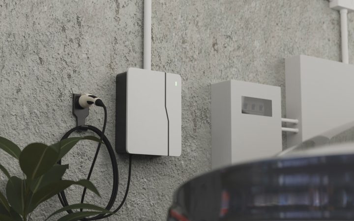 Modern white and black EV charger mounted on textured concrete wall with coiled black charging cable and connector, green LED indicator light visible, partial view of electric vehicle in foreground