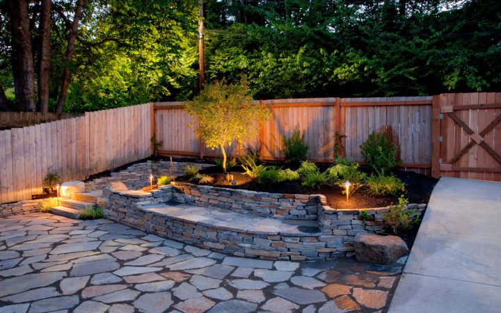 An elegantly landscaped backyard at twilight featuring a flagstone patio, a tiered stone retaining wall, and strategically placed low-voltage path lights illuminating the plants and walkway.