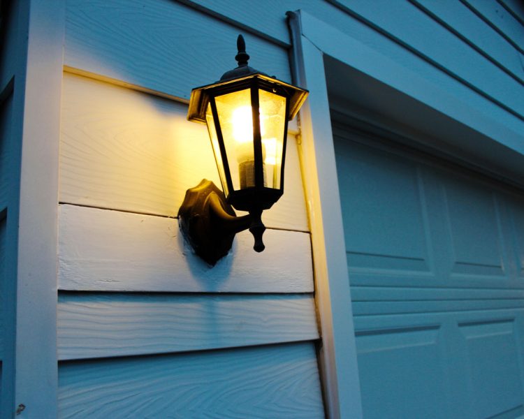 A traditional black metal and glass lantern-style light fixture mounted on white horizontal siding, casting a warm glow at dusk next to a white garage door.